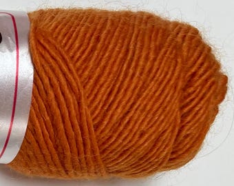 May include: Close-up of a ball of orange yarn. The yarn is tightly wound, showing individual strands with a slightly fuzzy texture. The color is a warm, vibrant orange, suitable for knitting or crochet projects. A white label with red accents is partially visible.
