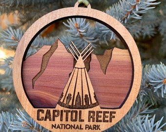 Puede incluir: Un adorno de madera con una imagen estilizada del Parque Nacional Capitol Reef, Utah. El adorno es circular y presenta una cordillera de color rojo con una tienda de campaña en el centro. El texto "CAPITOL REEF NATIONAL PARK" está impreso debajo de la imagen.
