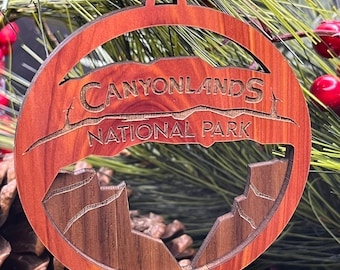 Puede incluir: Un adorno navideño de madera con un acabado de madera de grano marrón y rojo. El adorno presenta un recorte del horizonte del Parque Nacional Canyonlands y el texto "Canyonlands National Park".