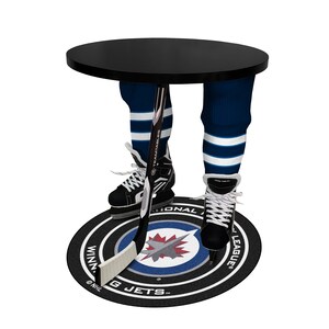 May include: A black circular table supported by hockey-themed legs. The legs are made of blue and white striped hockey socks, skates, and a hockey stick. The table stands on a Winnipeg Jets logo rug.