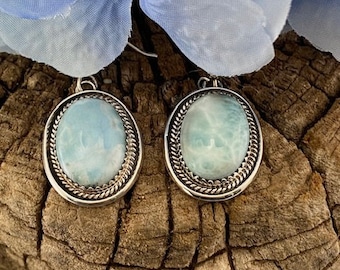 Könnte beinhalten: Ein Paar silberne Ohrringe mit ovalen, blauen und weißen Edelstein-Cabochons, die in einer silbernen Fassung mit einem dekorativen Rand gefasst sind.