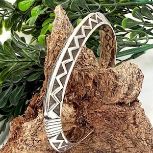 May include: A silver cuff bracelet with a geometric pattern of triangles and lines. The bracelet is displayed on a piece of textured wood, with green foliage in the background. The bracelet has a Native American design.