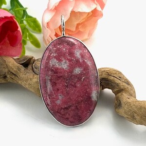 Peut inclure: Un pendentif en argent avec une pierre précieuse ovale rose et blanche. Le pendentif est placé sur un fond blanc avec un morceau de bois flotté et une fleur rose.