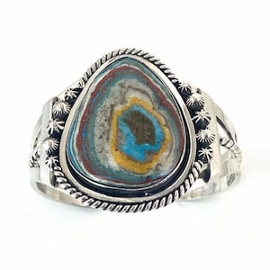 Puede incluir: Un brazalete de plata con una piedra grande, ovalada y multicolor en el centro. La piedra tiene tonos de azul, verde, amarillo, marrón y rojo.