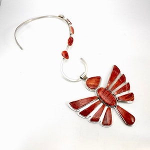 May include: A silver necklace with a large, red, spiny oyster shell pendant. The pendant is shaped like a stylised bird with outstretched wings. The necklace is made of a thin silver chain with several smaller red spiny oyster shell accents.