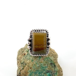May include: A silver ring with a rectangular brown and yellow gemstone set in a rope-like border.