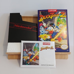 Peut inclure: Une cartouche de jeu et une boîte de jeu Nintendo Entertainment System pour DuckTales 2, un jeu vidéo mettant en scène le personnage Disney de Picsou. La boîte est bleue et présente une image de Picsou, Riri, Fifi et Loulou. La cartouche de jeu est noire et porte une étiquette avec le titre du jeu.