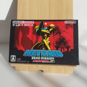 Puede incluir: Una caja de cartucho de juego Game Boy Advance roja, negra y amarilla con el título "Metroid Zero Mission" y el texto "ゲームボーイアドバンス" en japonés. La caja presenta un personaje con un traje amarillo sosteniendo un arma.
