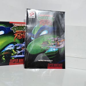 Könnte beinhalten: Super Nintendo-Spielkassette Teenage Mutant Ninja Turtles: Tournament Fighters mit schwarzer Box und Anleitung. Die Box zeigt eine grüne Teenage Mutant Ninja Turtle mit einem blauen Bandana. Die Anleitung zeigt eine Nahaufnahme der gleichen Schildkröte.