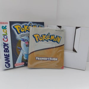 Puede incluir: Una caja de videojuego Game Boy Color y una Guía del Entrenador para Pokémon. La caja es azul y negra con el logo de Pokémon y una criatura blanca y azul. La Guía del Entrenador está en una funda de plástico transparente.