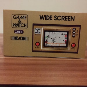 Op de afbeelding: Een gouden Game & Watch Wide Screen handheld-spelconsole met het spel "Chef" weergegeven op het scherm. De console heeft een rode knop aan de linker- en rechterkant en een zwart-wit scherm met een gepixelde afbeelding van een kok die kookt.