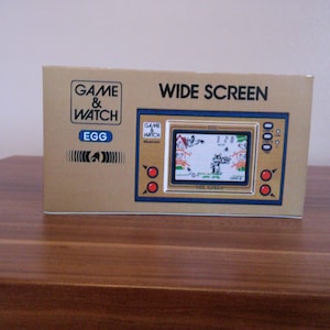 May include: A brown and gold Game & Watch handheld game console with a wide screen. The game is called "Egg" and features a cartoon chicken on the box art. The console is on a wooden surface.