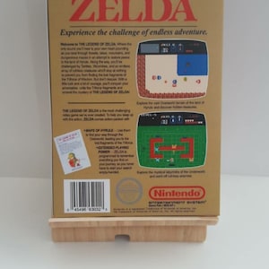 Może przedstawiać: Kaseta z grą The Legend of Zelda do systemu rozrywki Nintendo. Tył pudełka przedstawia złote tło z opisem gry i zrzutami ekranu. Gra jest opisana jako wymagająca przygoda z mapami, rozszerzoną mocą gry i r&oacute;żnymi wrogami.
