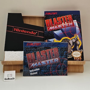 Könnte beinhalten: Ein schwarzes Nintendo-Spielkassettengehäuse mit dem Text "Nintendo®" auf der Vorderseite. Das Gehäuse befindet sich neben einer Videospiel-Anleitung mit dem Titel "BLASTER MASTER" in rot und blauem Text. Die Anleitung hat einen blauen und schwarzen Hintergrund mit einem roten und weißen Ziel-Design. Die Anleitung hat auch ein goldenes Siegel mit dem Text "Official Nintendo Seal" darauf.
