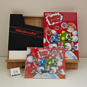 Könnte beinhalten: Eine rote und weiße Nintendo-Spielkassettenbox für das Spiel Bubble Bobble. Die Box zeigt ein Bild von zwei grünen und blauen Dinosauriern, die Blasen pusten. Der Text auf der Box lautet "Taito Bubble Bobble".