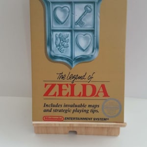 Może przedstawiać: Złote pudełko na kartridż gry Nintendo Entertainment System do gry The Legend of Zelda. Pudełko zawiera błękitno-biały herb z kluczem, sercami i lwem. Tekst "The Legend of Zelda" jest w kolorze czerwonym i białym.