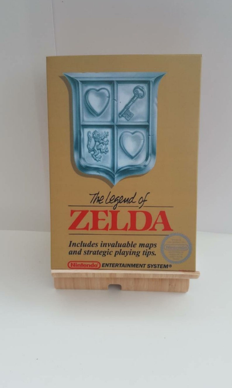 Może przedstawiać: Złote pudełko na kartridż gry Nintendo Entertainment System do gry The Legend of Zelda. Pudełko zawiera błękitno-biały herb z kluczem, sercami i lwem. Tekst "The Legend of Zelda" jest w kolorze czerwonym i białym.