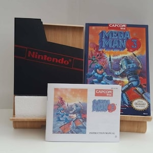 Könnte beinhalten: Ein komplettes Set des klassischen Nintendo Entertainment System-Spiels Mega Man 3, inklusive Spielkassette, Anleitung und Box. Die Box ist schwarz mit dem Nintendo-Logo in Rot. Die Spielkassette befindet sich in einem Plastikgehäuse mit einem blau-weißen Etikett, auf dem "CAPCOM USA MEGA MAN 3" steht. Die Anleitung ist weiß mit einem blau-weißen Etikett, auf dem "CAPCOM USA MEGA MAN 3" steht.