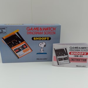 May include: A blue box with a yellow and black Game & Watch Panorama Screen Snoopy handheld video game. The box has the text "GAME & WATCH™ PANORAMA SCREEN SNOOPY" and "Nintendo®" on it. There is a white instruction booklet with the text "GAME & WATCH PANORAMA SCREEN SNOOPY (SM-91) INSTRUCTION Nintendo®" on it.