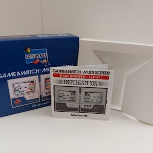 Puede incluir: Una caja blanca con una tapa azul y un folleto de instrucciones Game & Watch Multi Screen Rain Shower (LP-57). La caja tiene una imagen del Game & Watch en la parte delantera. El folleto de instrucciones tiene una imagen del Game & Watch en la parte delantera.