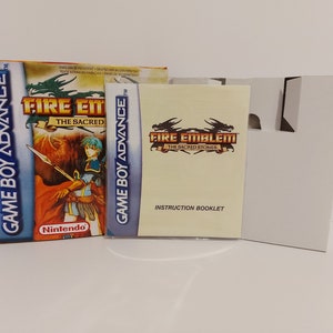 Könnte beinhalten: Eine Game Boy Advance-Spielkassette und eine Anleitung für Fire Emblem: The Sacred Stones. Das Cover zeigt einen Charakter, der eine Lanze hält, und einen Drachen im Hintergrund. Die Anleitung hat einen gelben Einband mit einem Drachen und dem Spieltitel.
