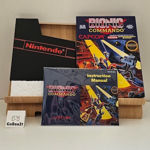 May include: A black Nintendo game cartridge case with the text "Nintendo" in red. The case is open and shows a black and white game cartridge with the text "Bionic Commando" and "Capcom" on it. The game is for the Nintendo Entertainment System.