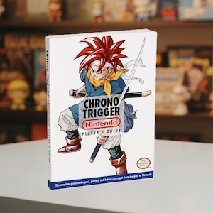 Könnte beinhalten: Ein weißes Softcover-Spielerhandbuch für Chrono Trigger von Nintendo mit einer zentralen Illustration einer Figur mit stacheligen roten Haaren, die ein Katana hält. Das Cover enthält den Spieletitel, das Nintendo-Logo und den Text: The complete guide to the past, present and future—straight from the pros at Nintendo.