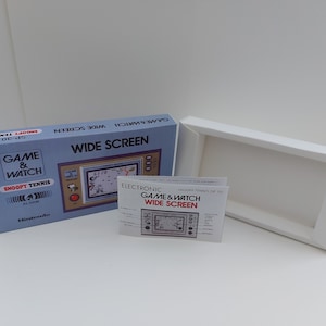 Puede incluir: Una consola de juegos portátil Nintendo Game & Watch Wide Screen azul y blanca con una imagen de Snoopy jugando al tenis en la pantalla. La caja está abierta y el manual de instrucciones es visible. El texto en la caja dice "Electronic Game & Watch Wide Screen" y "Snoopy Tennis SP-30".