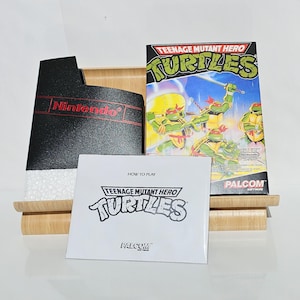 Peut inclure: Une cartouche de jeu vidéo Teenage Mutant Ninja Turtles pour la Nintendo Entertainment System. Le jeu est emballé dans un boîtier de cartouche noir avec un logo Nintendo rouge. La jaquette présente les quatre Tortues Ninja dans une pose de combat. La boîte est intitulée "Teenage Mutant Hero Turtles" et "PALCOM Software".