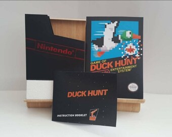 May include: A black Nintendo Entertainment System game cartridge with a black instruction booklet and a blue and green box art featuring a duck flying with a red target. The box art reads "Game of Duck Hunt".