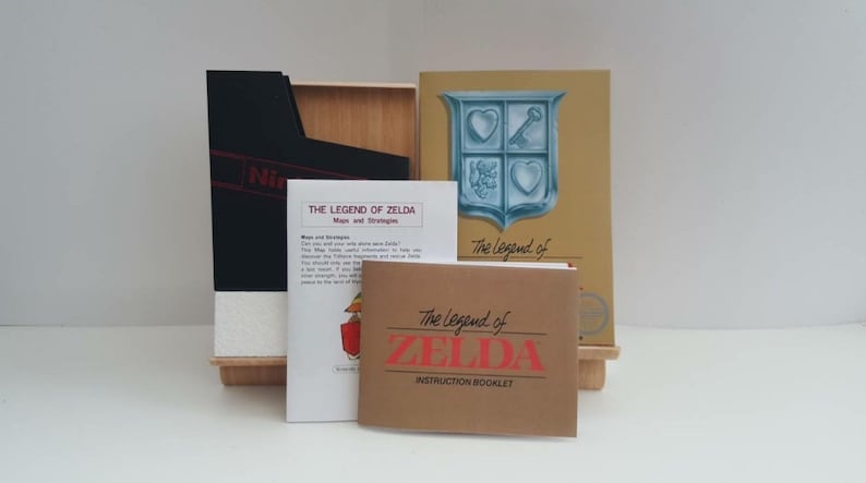 Może przedstawiać: Oryginalna kaseta do gry Nintendo Entertainment System (NES) z grą The Legend of Zelda. Gra znajduje się w oryginalnym pudełku, kt&oacute;re jest czarne z czerwonym logo Nintendo. Pudełko zawiera r&oacute;wnież instrukcję gry oraz mapę i przewodnik po strategii.
