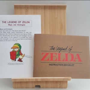 Może przedstawiać: Brązowa i biała instrukcja do gry wideo "The Legend of Zelda" z tytułem "Instruction Booklet" wydrukowanym na okładce. Instrukcja leży na drewnianym stojaku obok mniejszej białej instrukcji z tytułem "The Legend of Zelda: Maps and Strategies".