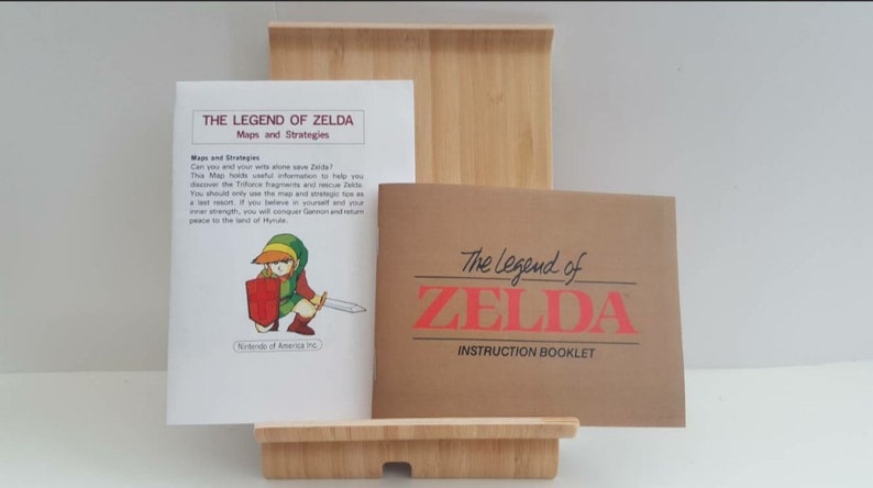 Może przedstawiać: Brązowa i biała instrukcja do gry wideo "The Legend of Zelda" z tytułem "Instruction Booklet" wydrukowanym na okładce. Instrukcja leży na drewnianym stojaku obok mniejszej białej instrukcji z tytułem "The Legend of Zelda: Maps and Strategies".