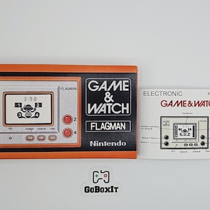 Op de afbeelding: Een vintage Nintendo Game & Watch "Flagman" handheld elektronisch spel. Het spel heeft een zwart en oranje frame met de speltitel en het logo. Het scherm toont een spel in uitvoering. Inclusief de originele handleiding.
