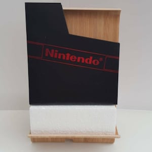 Może przedstawiać: Czarny wkład do gier Nintendo z czerwonym logo Nintendo na drewnianym stojaku. Wkład leży na białej podstawie z pianki.