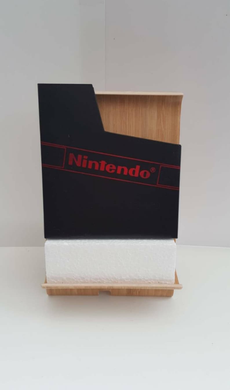 Może przedstawiać: Czarny wkład do gier Nintendo z czerwonym logo Nintendo na drewnianym stojaku. Wkład leży na białej podstawie z pianki.