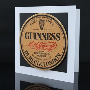 May include: A greeting card featuring a vintage Guinness stout beer coaster design. The coaster is circular, with the Guinness logo and text in black and red on a tan background. The card is white with a black border.