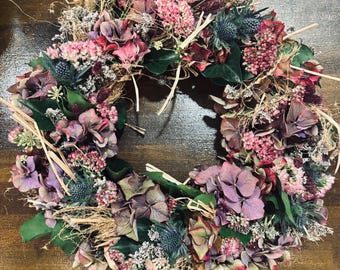 Könnte beinhalten: Ein kreisförmiger Kranz aus getrockneten Blumen in Rosa-, Lila- und Grüntönen. Der Kranz besteht aus Hortensien, Disteln und anderen getrockneten floralen Elementen mit Strohakzenten. Die Anordnung befindet sich auf einer dunklen Holzoberfläche.
