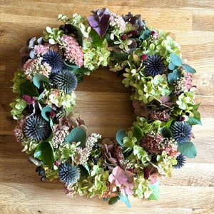 Könnte beinhalten: Ein dekorativer, kreisförmiger Kranz aus getrockneten Blumen und Blättern. Der Kranz zeigt eine Mischung aus Farben, darunter Grün, Rosa, Lila und Blau. Er enthält Hortensien, Disteln und andere Elemente und bietet ein natürliches, rustikales Aussehen.