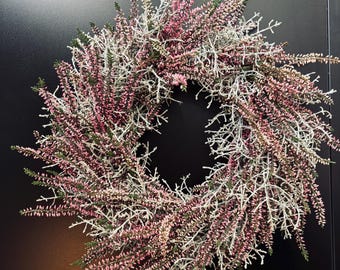 Könnte beinhalten: Ein kreisförmiger Kranz aus getrockneten rosa, grünen und weißen Blumen und Zweigen. Der Kranz ist vor einem schwarzen Hintergrund an einer rosa Schnur aufgehängt. Die Blumenanordnung ist dicht und voll und schafft ein natürliches, dekoratives Element.
