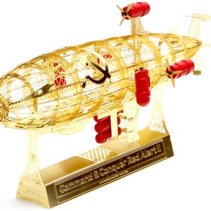 May include: A gold metal model of a Soviet airship from the video game Command & Conquer: Red Alert 2. The airship has a red star and hammer and sickle symbol on the side. The model is on a stand with the game's title on it.