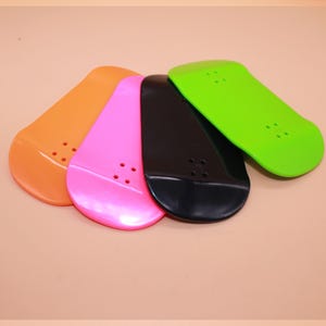 Könnte beinhalten: Vier farbenfrohe Finger-Skateboards in Orange, Pink, Schwarz und Grün. Jedes Board hat eine abgerundete Form mit vier kleinen Löchern. Die Boards sind fächerförmig angeordnet und zeigen ihre leuchtenden Farben.