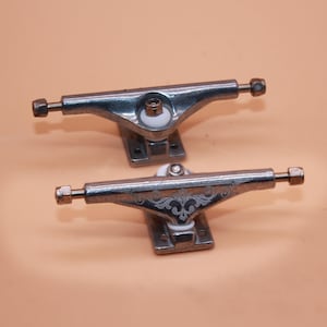 Puede incluir: Dos ejes de skate de plata con bujes blancos. Los ejes tienen un diseño decorativo grabado en el metal.
