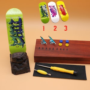 Puede incluir: Un conjunto de tres cubiertas de fingerboard con diferentes diseños gráficos, una rampa de madera con tornillos de metal, dos ejes de metal azul, cuatro ruedas amarillas, un destornillador y una alfombrilla de herramientas negra. Las fingerboards están etiquetadas con 1, 2 y 3.