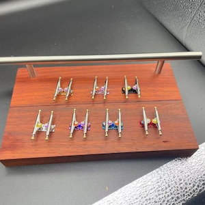May include: A wooden display stand with a silver metal bar on top. The stand has two levels and is filled with 10 sets of fingerboard trucks. The trucks are silver with gold accents and have different colored wheels.
