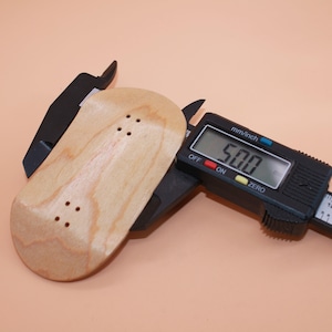 Puede incluir: Una tabla de dedo de madera de color claro se mide con un calibrador digital. El calibrador muestra una medida de 5,00 mm. La tabla de dedo tiene cuatro agujeros pretaladrados para el montaje.
