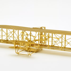 Op de afbeelding: Een gouden metalen model van de Wright Flyer van de gebroeders Wright uit 1903. Het model is gedetailleerd en bevat de vleugels, propellers en het landingsgestel.