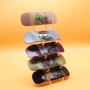 Peut inclure: Un ensemble de cinq fingerboards avec des motifs graphiques colorés. Les fingerboards sont exposés sur un support en acrylique transparent. Les motifs comprennent une créature verte, une créature noire et blanche, une créature violette, une créature verte et blanche et une créature bleue et blanche.