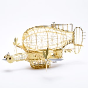 Puede incluir: Un modelo de dirigible de metal dorado con un gran globo redondo de estructura abierta y una góndola rectangular más pequeña de estructura abierta. El dirigible tiene múltiples hélices y una nariz puntiaguda.