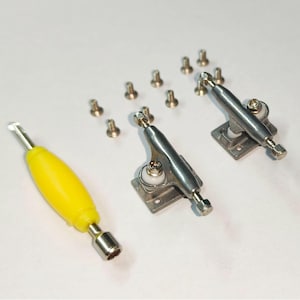 Peut inclure: Ensemble de pièces de skateboard miniatures, comprenant deux trucks en métal argenté, un outil jaune et plusieurs petites vis argentées. Les composants sont disposés sur une surface blanche, suggérant un kit de bricolage.